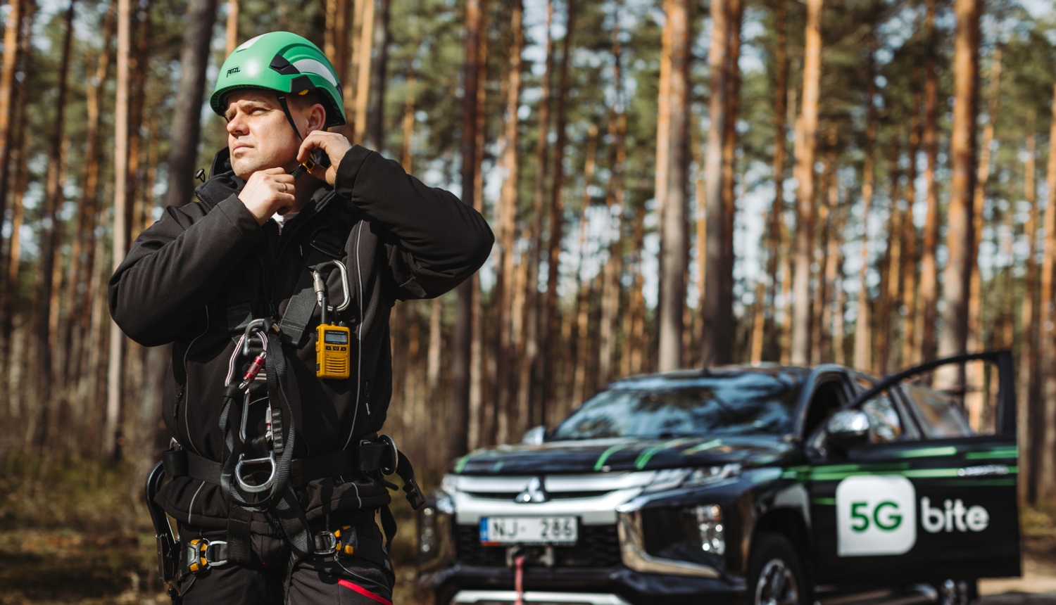 "Bite" telekomunikāciju inženieris darba ekipējumā un zaļā aizsargķiverē gatavojas darbam meža apvidū. Fonā redzams apvidus auto ar "5G bite" zīmolu.