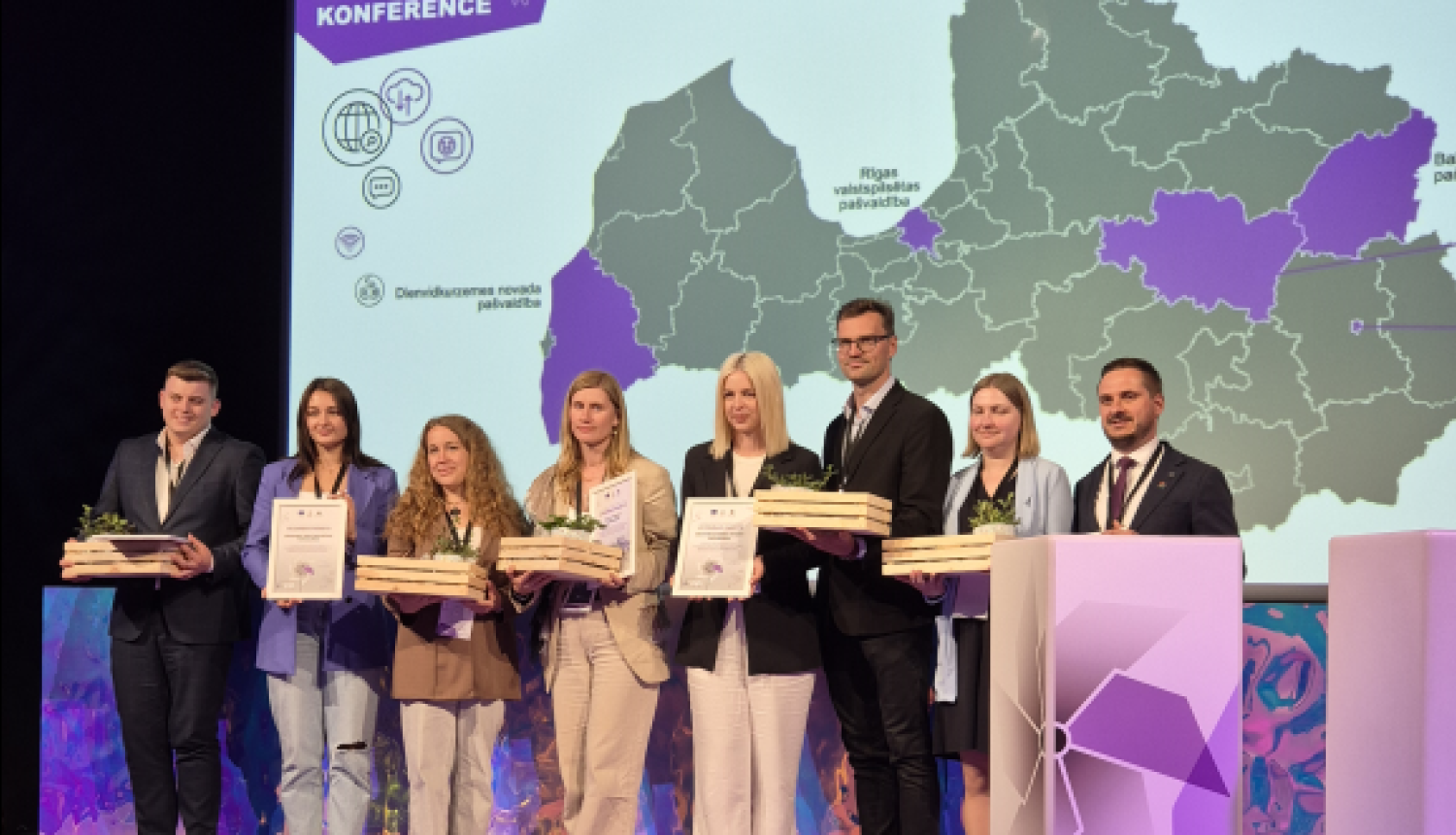 Projekta "Digitālā darba ar jaunatni sistēmas attīstība pašvaldībās" noslēguma konferences dalībnieki uz skatuves ar apbalvojumiem un diplomiem rokās. Fonā ekrāns ar Latvijas karti un uzrakstu "SADARBĪBAS MEISTARI".