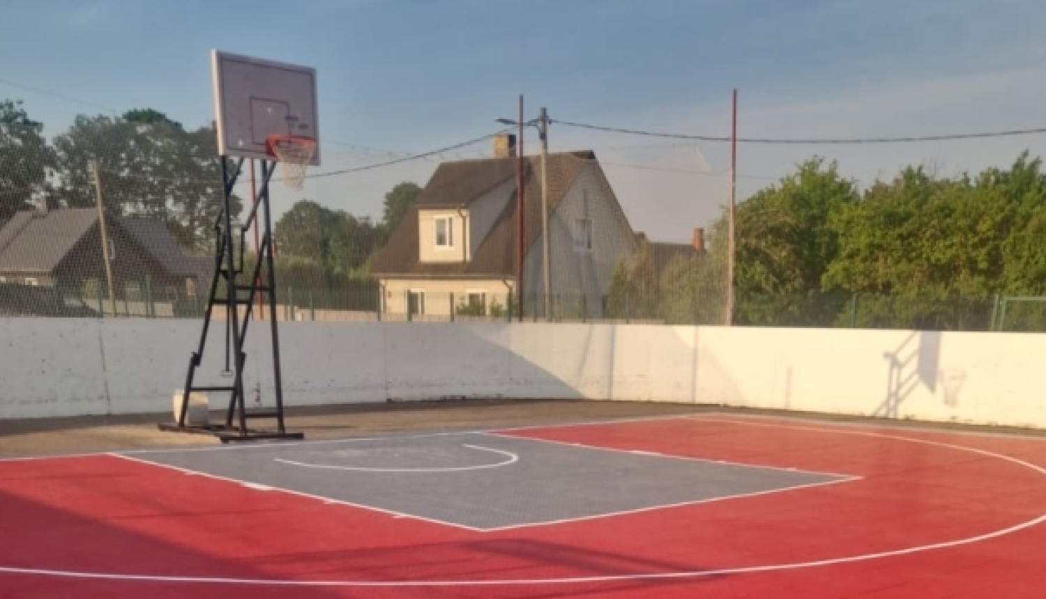 Āra basketbola laukums ar spilgti sarkanu un pelēku moduļu segumu. Laukumā redzams basketbola grozs, un to ieskauj balta sēta.
