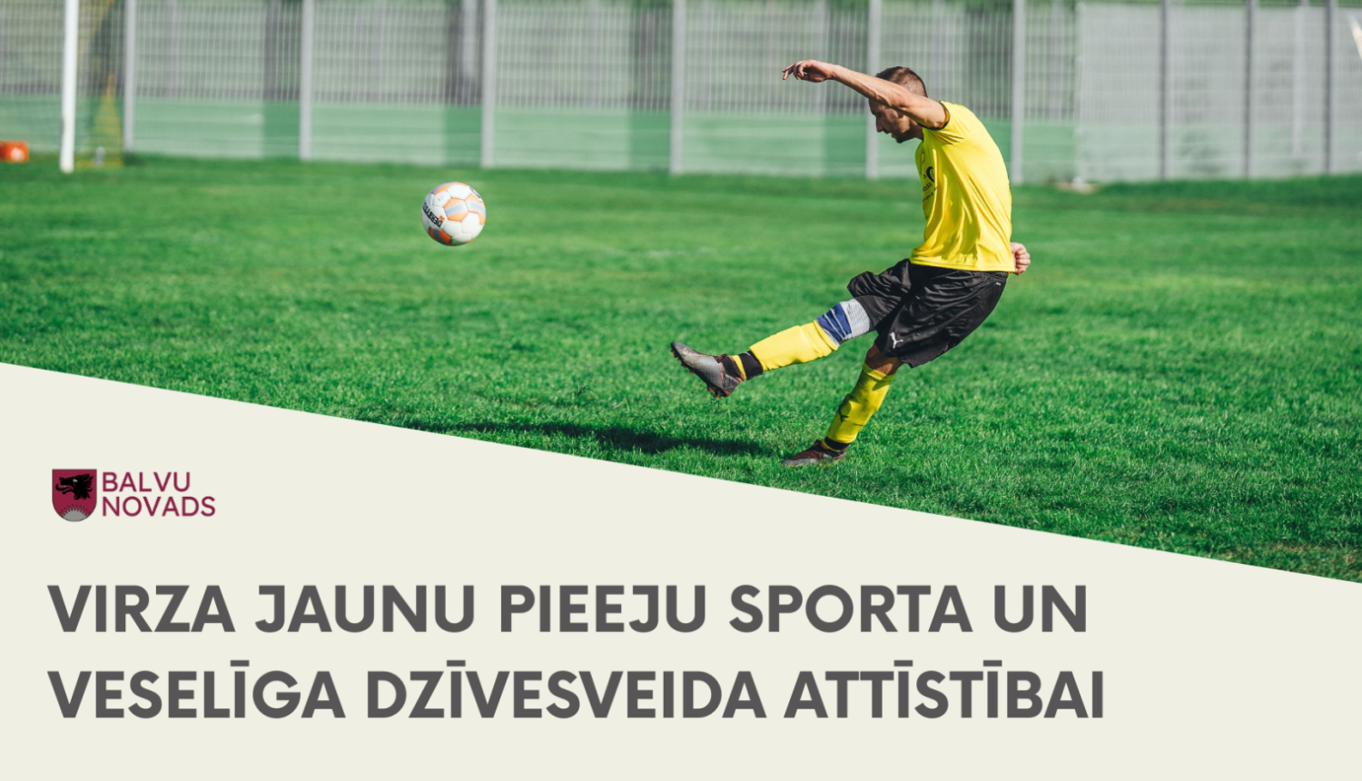 Futbolists dzeltenā t-kreklā un melnās šortos spēles gaitā sper pa futbola bumbu uz zaļa zālāja laukuma. Fonā redzams nožogojums un vārti.