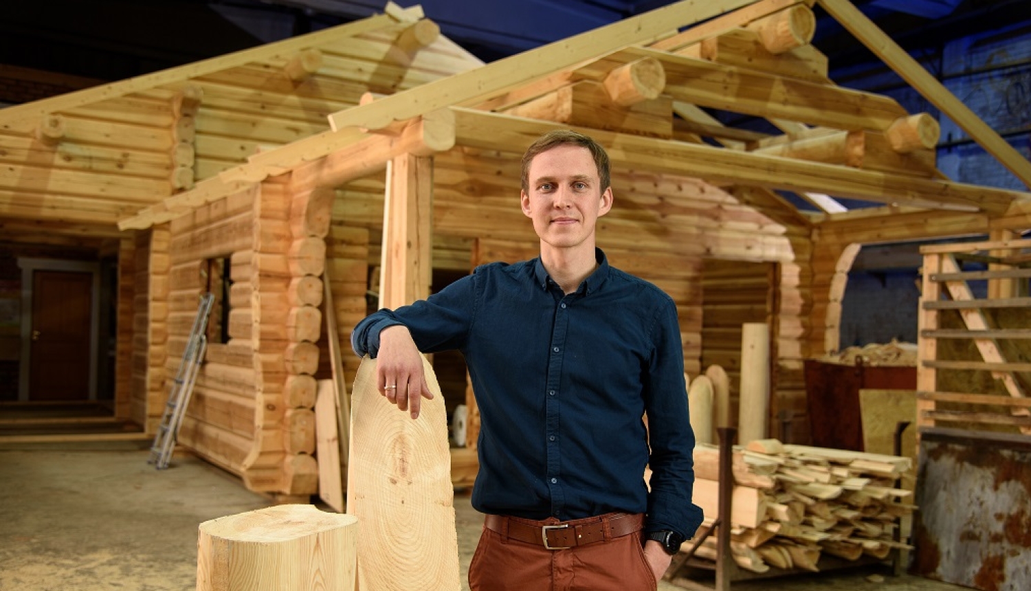Jānis Igaunis tumši zilā kreklā pozē ražošanas angārā uz nepabeigtas guļbūves koka mājas karkasa fona, atbalstījis roku pret lielu koka brusu.
