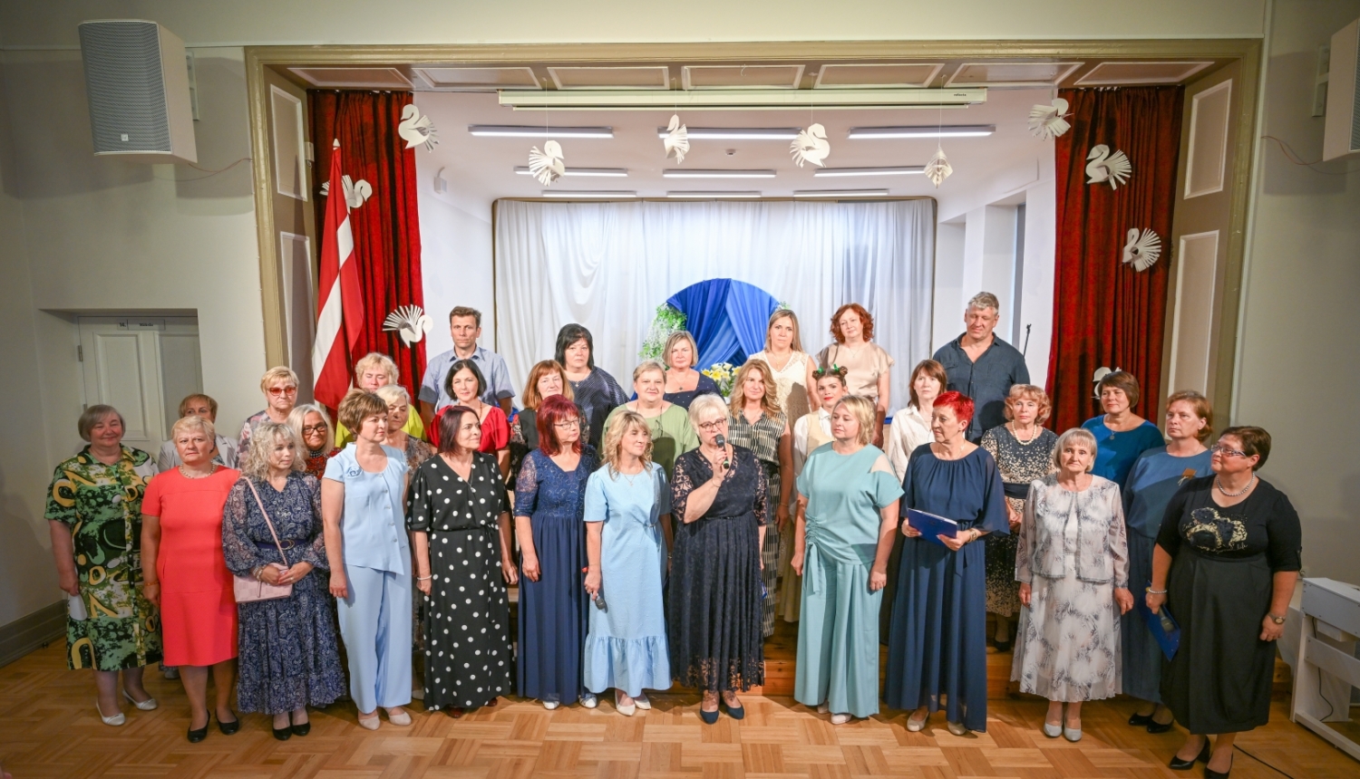 Attēlā redzama liela cilvēku grupa, kas stāv uz skatuves svinīgā pasākumā. Priekšplānā vairākas sievietes ir saposušās svētku tērpos, viena no viņām runā mikrofonā, bet cita tur zilu mapi. Fonā redzami sarkani aizkari, balti dekorējumi un Latvijas karogs, kas rada oficiālu un svinīgu noskaņu. Visi ir pavērsti pret skatītājiem, iespējams, kopīgai uzstāšanās reizei vai piemiņas foto.