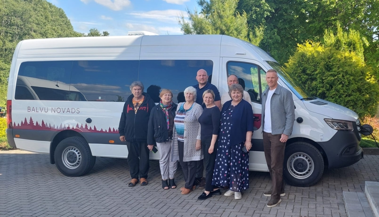 Cilvēku grupa pozē kopbildei pie balta mikroautobusa, uz kura sāniem ir uzraksts "BALVU NOVADS" un sarkans meža silueta krāsojums. Transportlīdzeklis ir pieslēgts uzlādes stacijai pie ķieģeļu ēkas.