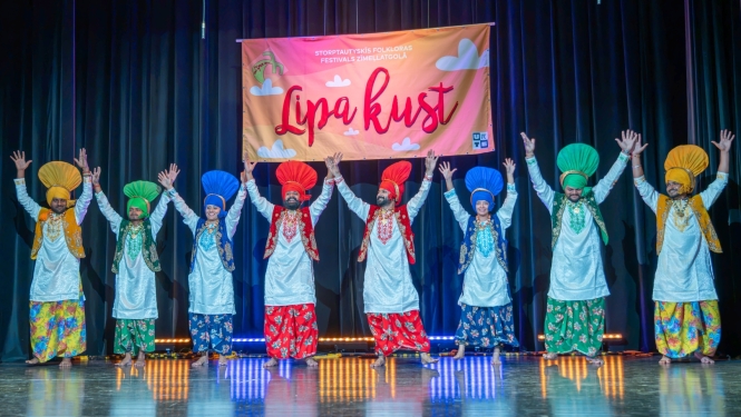 Uz skatuves rindā stāv astoņi tautastērpos ģērbušies dejotāji ar paceltām rokām. Viņi valkā košus, krāsainus tērpus un lielas tradicionālas galvassegas, radot svinīgu un enerģisku noskaņu. Fonā redzams festivāla plakāts un tumši skatuves aizkari, bet grīdu izgaismo skatuves apgaismojums.