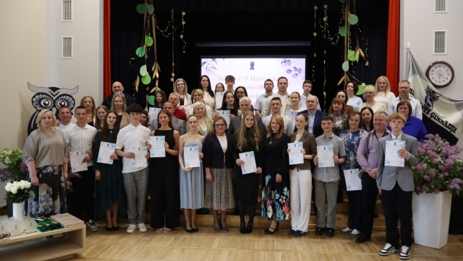 Attēlā redzama liela cilvēku grupa skolas vai ģimnāzijas aktu zālē. Daudzi jaunieši tur rokās diplomus vai apliecības, kas liecina par izlaidumu vai apbalvošanas pasākumu. Fonā ir skatuve ar dekorācijām, bet priekšplānā redzami ziedi un pedagogi, vecāki vai viesi, kas piedalās svinīgajā notikumā.