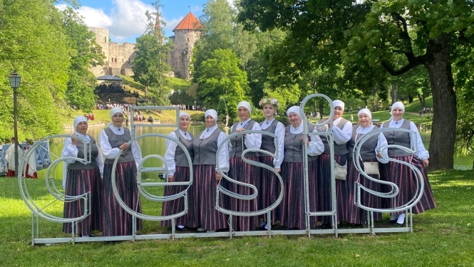 Sieviešu koris tautas tērpos pozē Cēsu Pils parka zālienā aiz liela, balta vides objekta — burtiem "CĒSIS". Fonā redzams dīķis un Cēsu viduslaiku pils drupas zaļumu ieskautā kalnā.