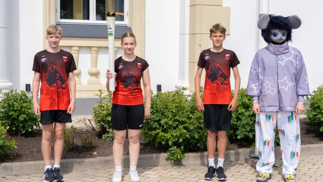 Attēlā redzami četri cilvēki, kas stāv ārā pie ēkas. Trīs jaunieši ir sporta tērpos sarkanmelnos kreklos, un viens no viņiem rokās tur degošu lāpu. Blakus viņiem stāv cilvēks pelēkā peles vai līdzīga dzīvnieka talismana kostīmā ar lielām ausīm un zilām acīm. Fonā redzama gaiša ēka ar logiem un apstādījumiem.