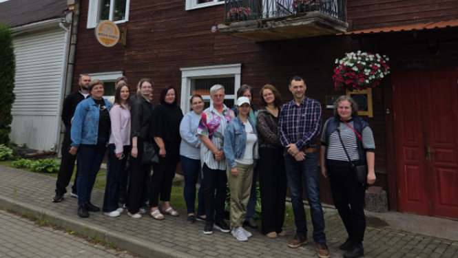 Cilvēku grupa pozē kopbildei uz ietves pie divstāvu koka ēkas ar balkonu un sarkanām ārdurvīm. Pie ēkas fasādes redzama apaļa izkārtne ar uzrakstu "Retro viesnīca".