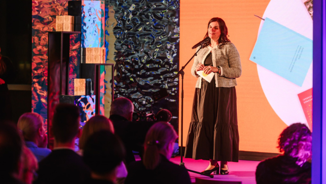 Attēlā redzama sieviete, kas stāv uz skatuves un runā mikrofonā auditorijas priekšā. Aiz viņas ir liels ekrāns ar oranžu fonu, uz kura redzams teksts "Zinātniskā literatūra". Priekšplānā redzami cilvēku silueti.