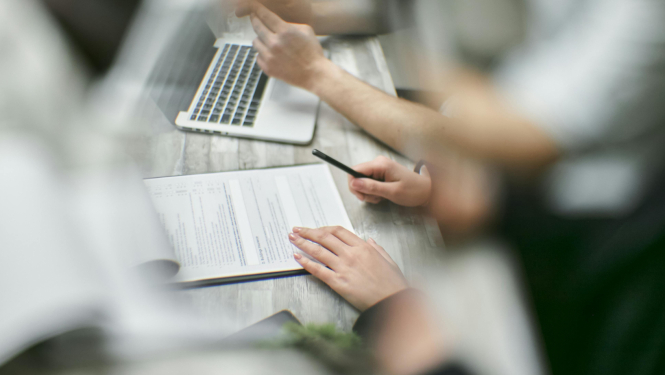 Divi cilvēki birojā strādā ar dokumentiem – viens tur rokās pildspalvu, otrs raksta pie atvērta dokumenta, fonā redzams klēpjdators.