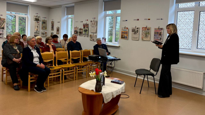 Gaišā telpā notiek neliels kultūras vai literārs pasākums. Sieviete stāv priekšā un lasa vai runā auditorijai, kamēr vairāki cilvēki sēž un klausās. Telpā redzams projektors, galds ar ziediem un uz sienām izkārtas avīžu pirmās lapas.