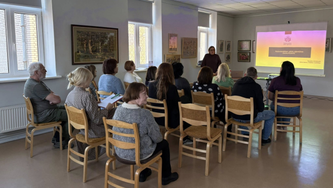 Gaišā telpā notiek informatīvs seminārs. Priekšā stāv lektore un runā pie projektora ekrāna, uz kura redzama prezentācija ar krāsainu fonu. Apmēram desmit klausītāji sēž rindās uz koka krēsliem, vērsti pret lektori; daži tur rokās pierakstu blokus un klausās.