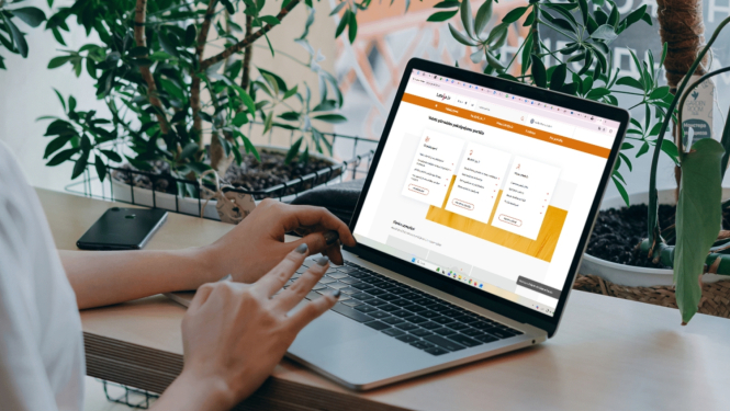 Persona sēž pie galda un lieto portatīvo datoru, kura ekrānā atvērta tīmekļvietne; fonā redzami zaļi telpaugi