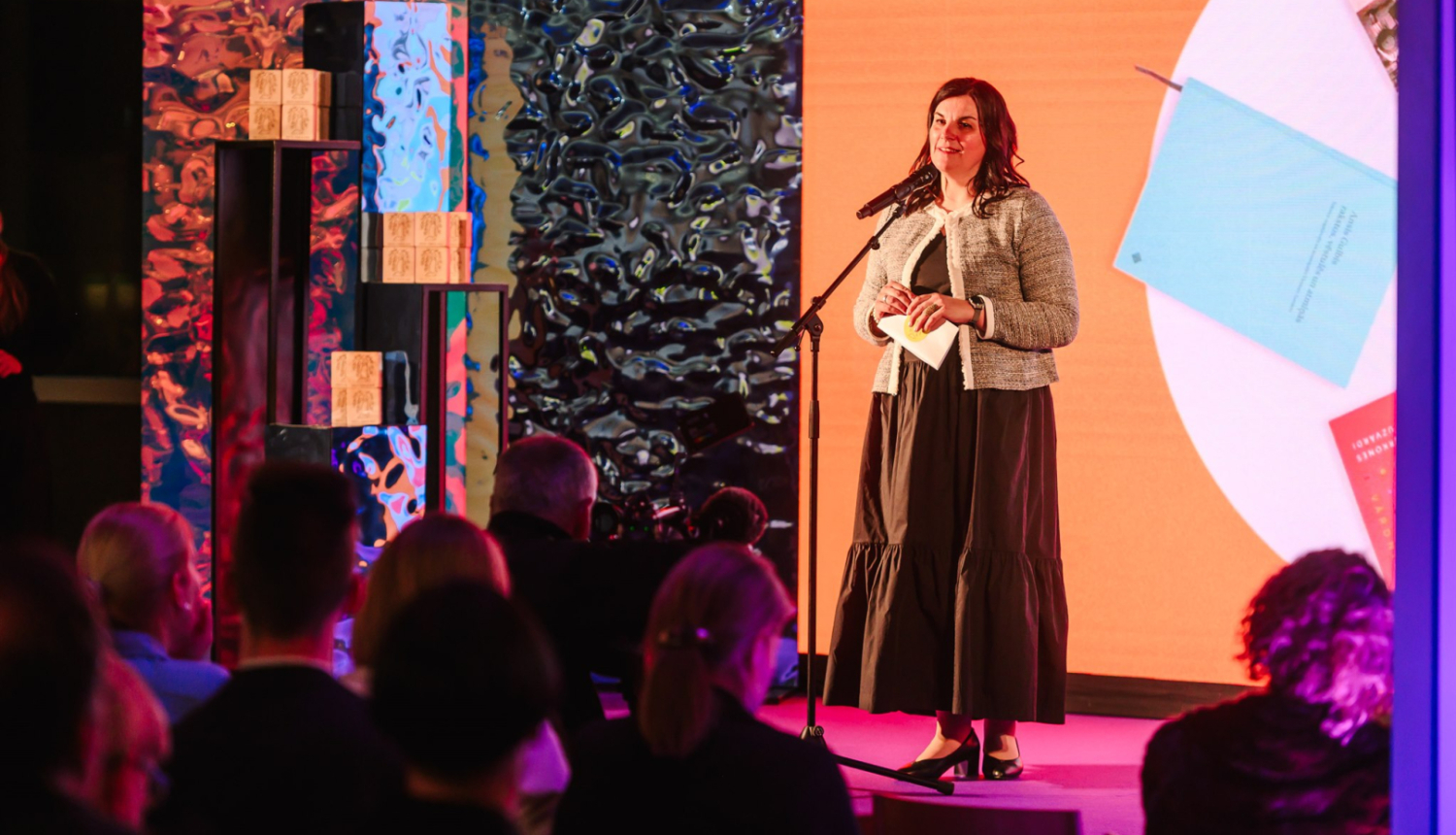 Attēlā redzama sieviete, kas stāv uz skatuves un runā mikrofonā auditorijas priekšā. Aiz viņas ir liels ekrāns ar oranžu fonu, uz kura redzams teksts "Zinātniskā literatūra". Priekšplānā redzami cilvēku silueti.