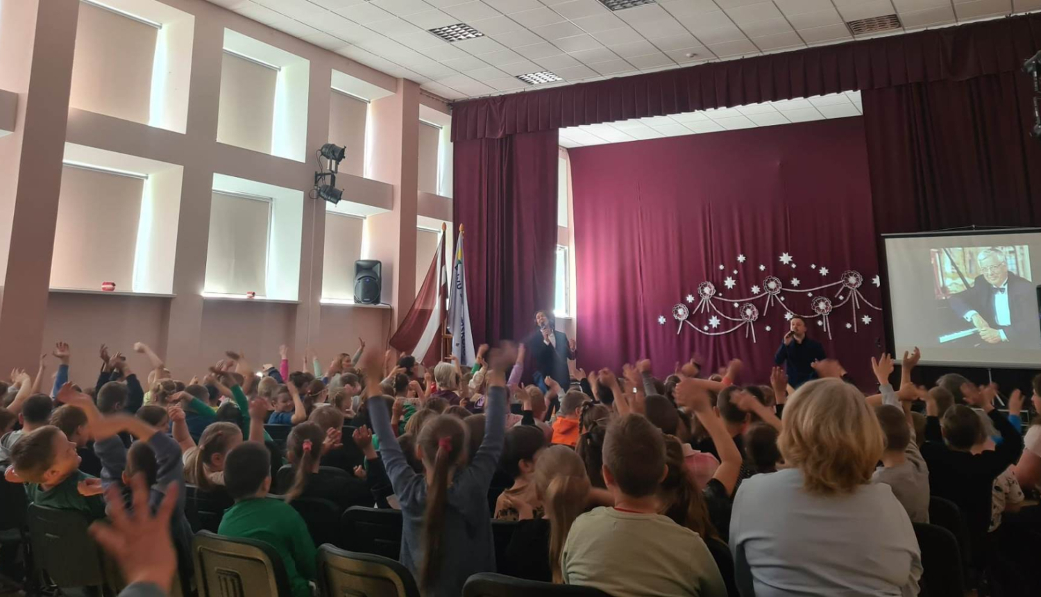 Attēlā redzama aktu zāle ar daudziem skolēniem. Skatuvi rotā bordo krāsas aizkari, un tās priekšā stāv divi vīrieši. Labajā pusē redzams ekrāns uz kura tiek projecēta Raimonda Paula bilde.