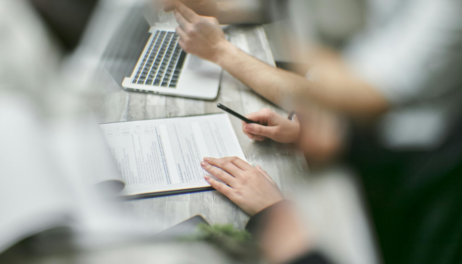 Divi cilvēki birojā strādā ar dokumentiem – viens tur rokās pildspalvu, otrs raksta pie atvērta dokumenta, fonā redzams klēpjdators.