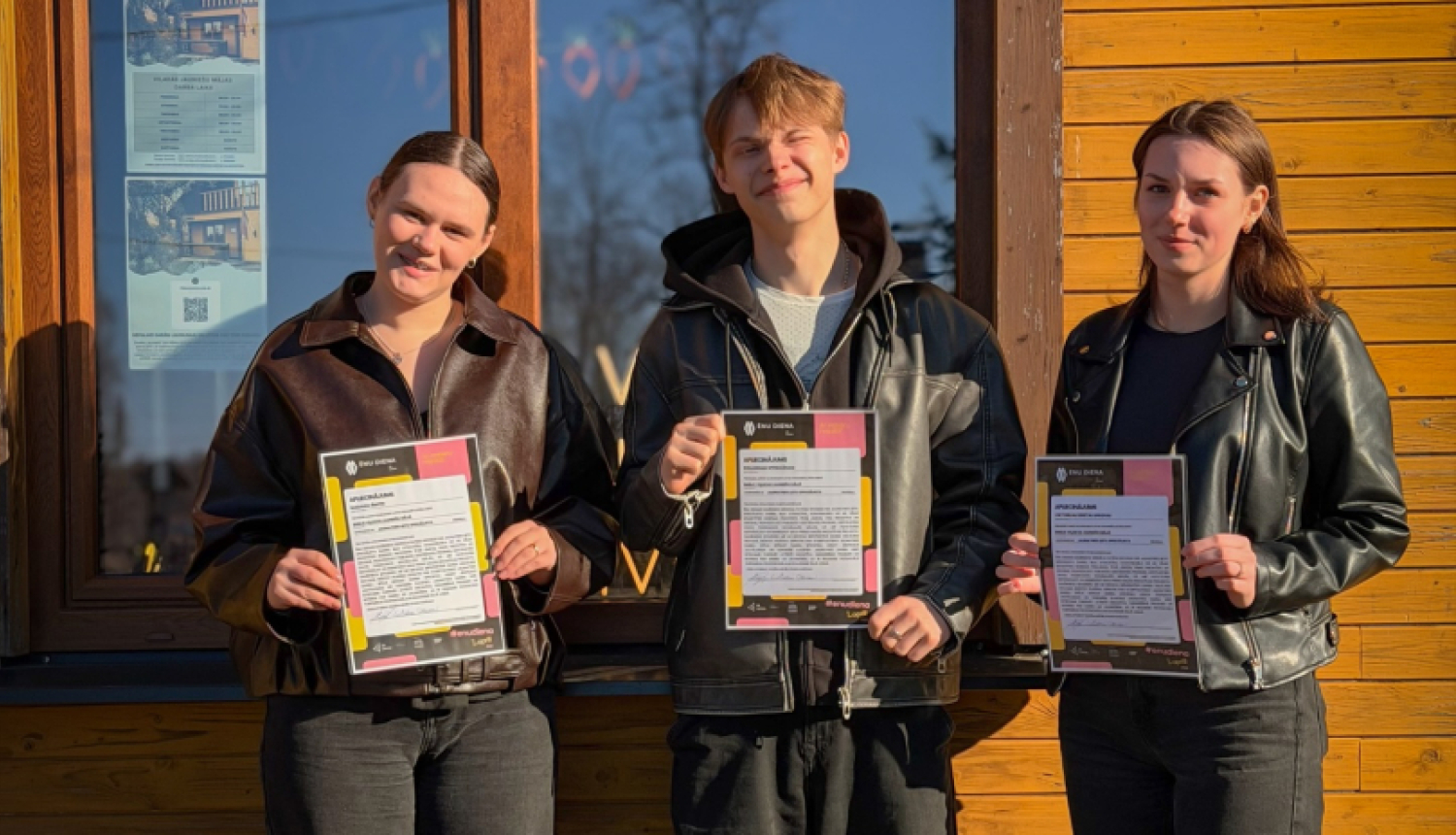 Trīs jaunieši ar sertifikātiem rokās stāv pie Viļakas jauniešu mājas