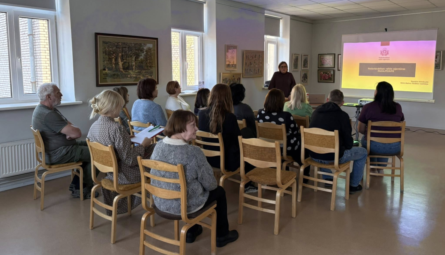 Gaišā telpā notiek informatīvs seminārs. Priekšā stāv lektore un runā pie projektora ekrāna, uz kura redzama prezentācija ar krāsainu fonu. Apmēram desmit klausītāji sēž rindās uz koka krēsliem, vērsti pret lektori; daži tur rokās pierakstu blokus un klausās.