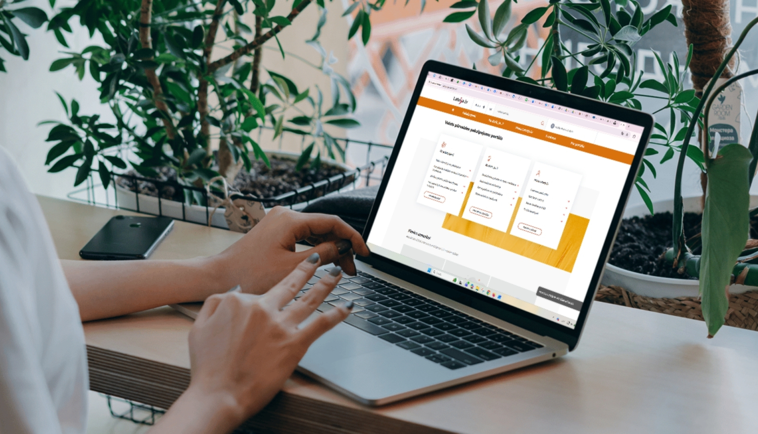 Persona sēž pie galda un lieto portatīvo datoru, kura ekrānā atvērta tīmekļvietne; fonā redzami zaļi telpaugi